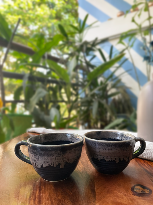 Pair of medium ceramic cups with handle, stoneware dark blue & grey glaze, on wooden table with greenery. Buy handcrafted mugs Bangalore.