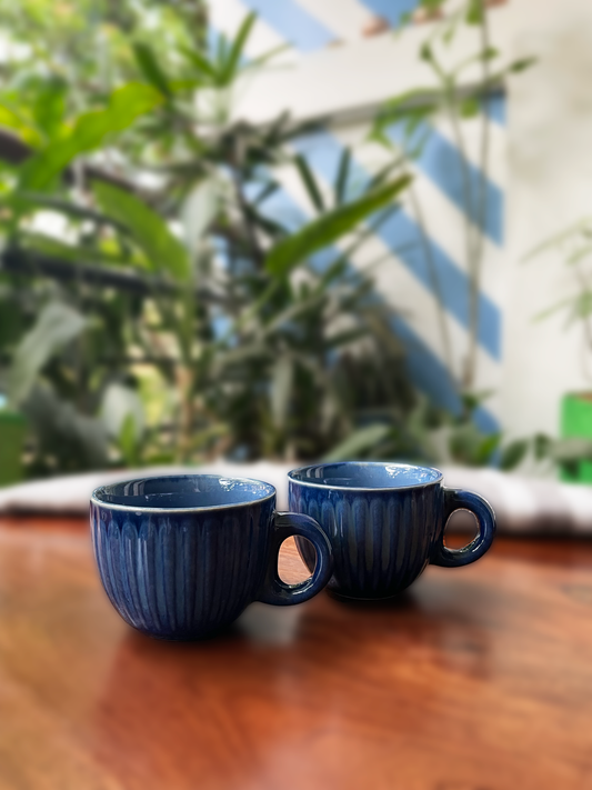 Pair of cute small ceramic cups with handle in shades of blue glaze, on table with blurry greenery background. Buy ceramics for Indian homes.